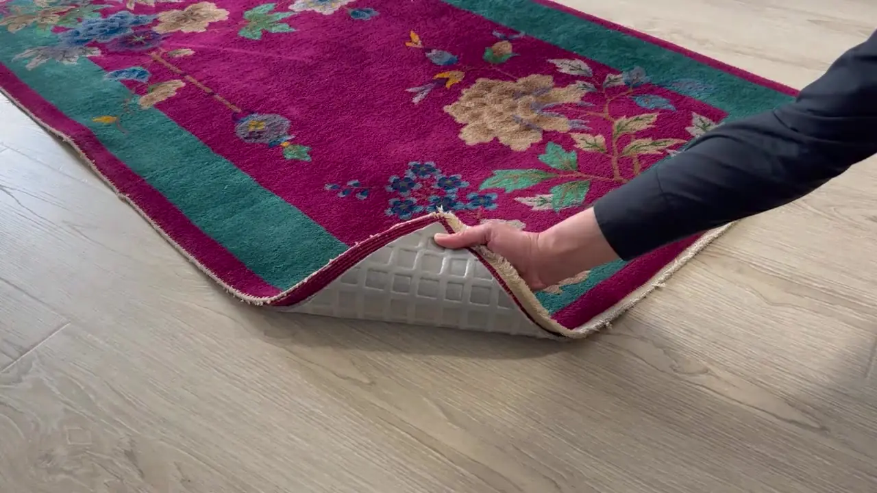 A hand lifting the corner of a magenta Oriental rug to show the Rug Pad Wizard felt pad underneath on a hardwood floor
